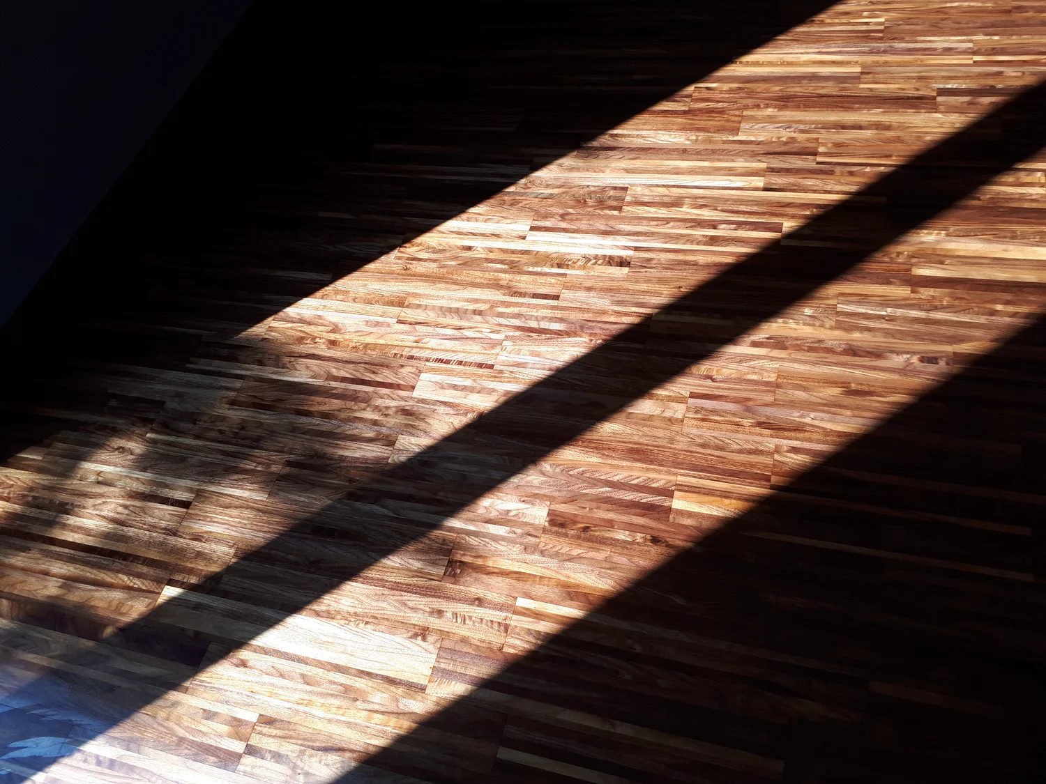 Schreinerei - Maßgefertigte Holzarbeiten - Ein Holzboden mit einer Mischung aus hellen und dunklen Holzdielen. Ein starker diagonaler Schatten fällt über den Boden, während helles Sonnenlicht die obere linke und die untere rechte Ecke erhellt.