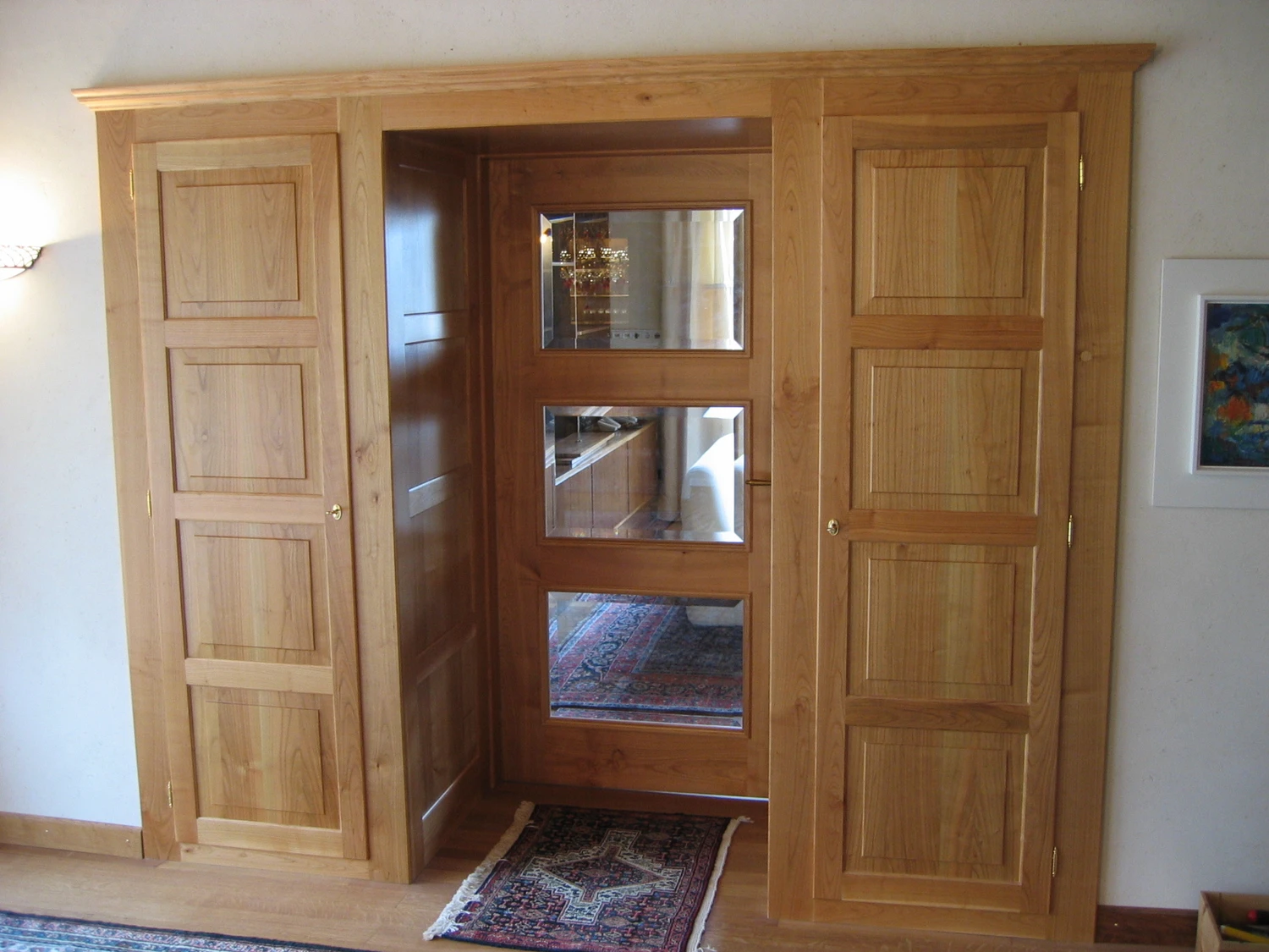 Schreinerei - Maßgefertigte Holzarbeiten - Innenansicht eines Zimmers mit hellen Holzschränken und einer Holztür. Die Tür besteht aus drei quadratischen Glasscheiben, die einen gemusterten Teppich und einen Teil des Raumes spiegeln. Die Schränke zu beiden Seiten der Tür sind ebenfalls mit Paneelen versehen.