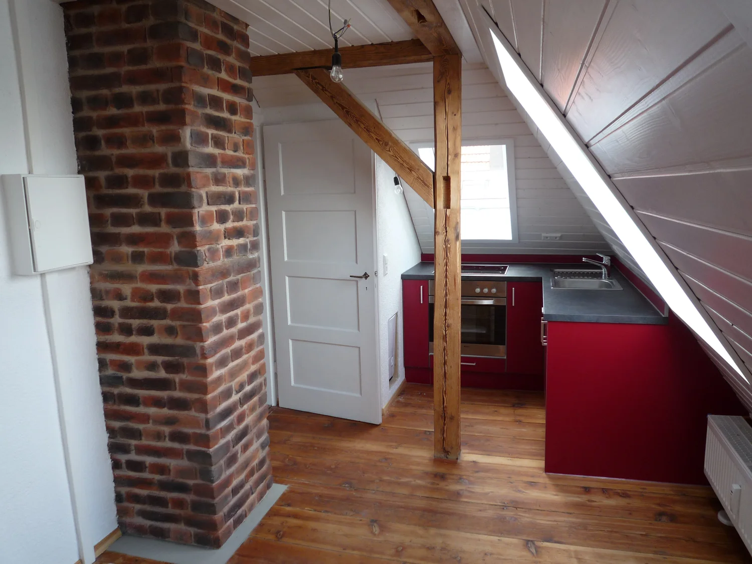 Schreinerei - Maßgefertigte Holzarbeiten - Eine kleine Dachgeschosswohnung mit Küche, roten Schränken, einer Backsteinsäule, einer weißen Tür und freiliegenden Holzbalken und -dielen.