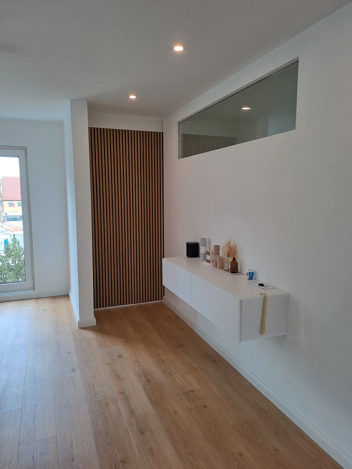 Schreinerei - Maßgefertigte Holzarbeiten - Innenansicht eines modernen Flurs mit hellem Holzboden, weißen Wänden, einer vertikalen Holzlamellenwand, einem weißen, schwebenden Konsolentisch und einem großen horizontalen Fenster darüber.