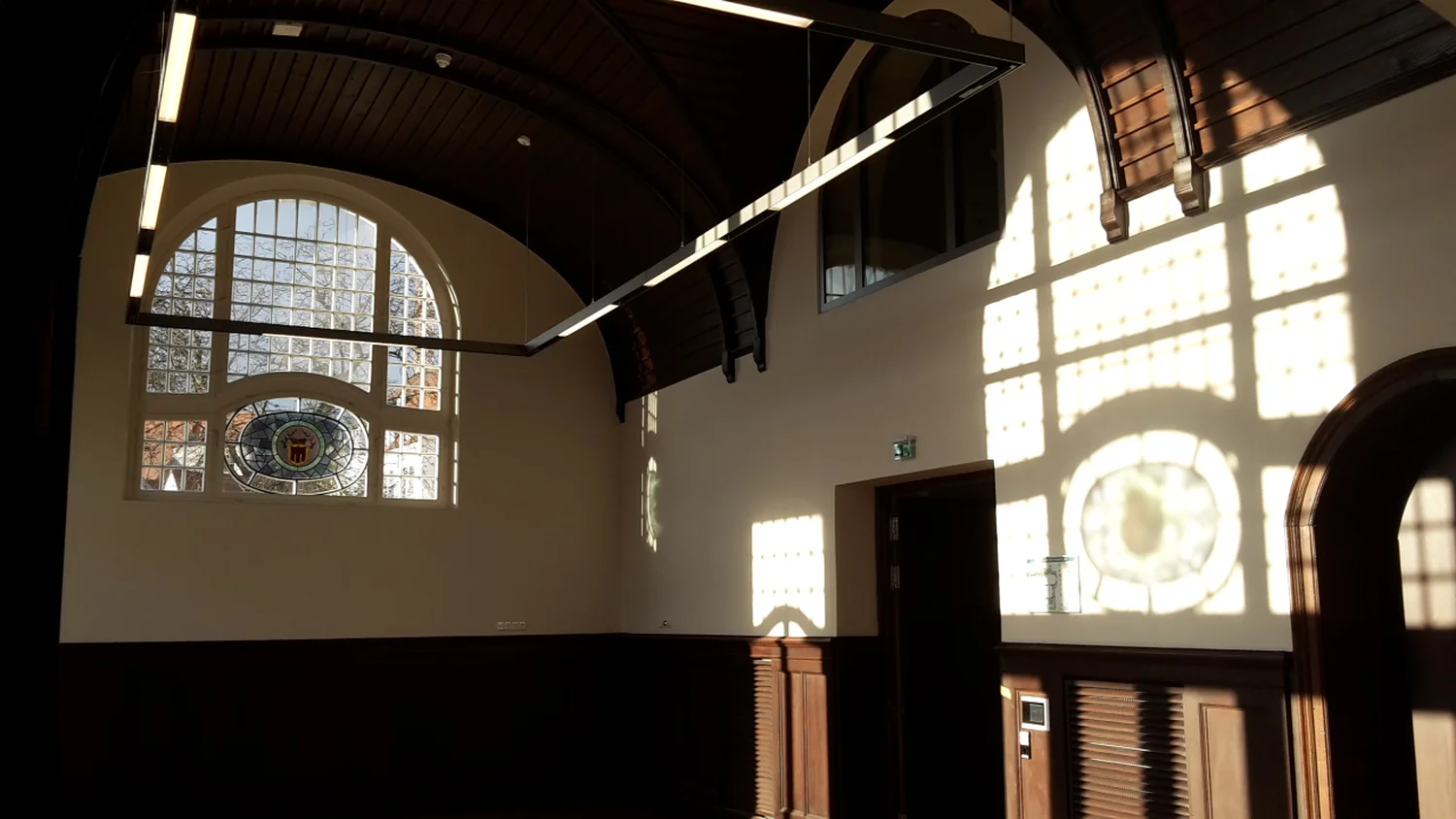 Schreinerei - Maßgefertigte Holzarbeiten - Das Innere der Kirche oder Halle zeichnet sich durch eine hohe, dunkle Holzgewölbedecke, weiße Wände, ein gewölbtes Buntglasfenster mit Sicherheitsgittern und durch die Fenster strömendes Sonnenlicht aus, das gemusterte Schatten auf Wände und Boden wirft.