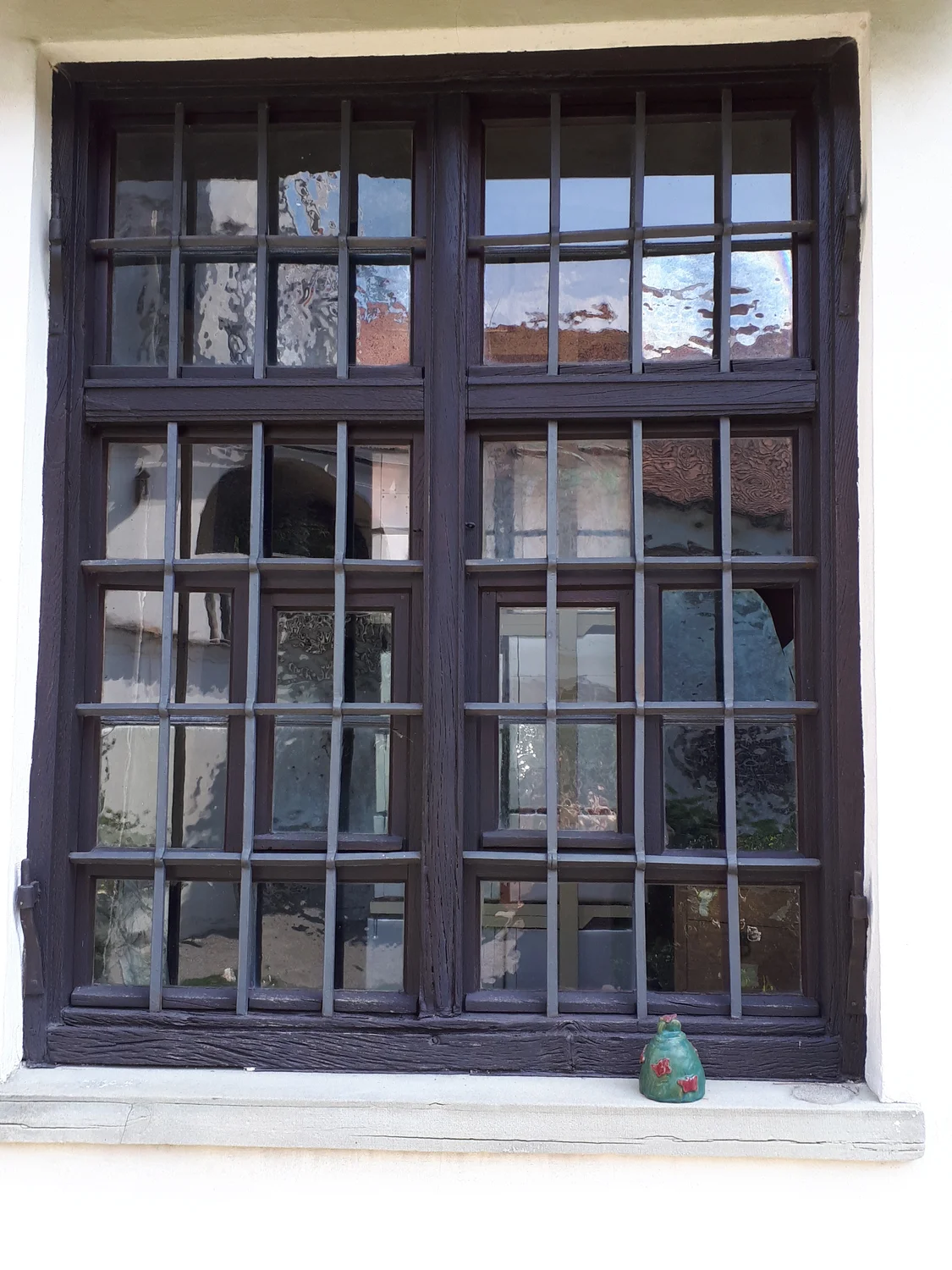 Schreinerei - Maßgefertigte Holzarbeiten - Ein altes, dunkles Holzfenster mit mehreren Scheiben und äußeren Metallgittern bietet Blick auf ein anderes Gebäude. Auf dem weißen Fensterbrett sitzt eine kleine grüne Froschfigur.