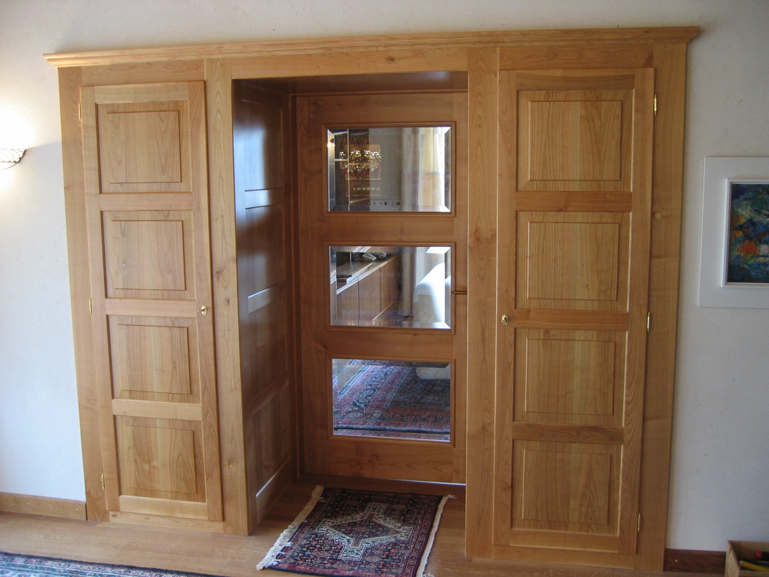 Schreinerei - Maßgefertigte Holzarbeiten - Innenansicht eines Zimmers mit hellen Holzschränken und einer Holztür. Die Tür besteht aus drei quadratischen Glasscheiben, die einen gemusterten Teppich und einen Teil des Raumes spiegeln. Die Schränke zu beiden Seiten der Tür sind ebenfalls mit Paneelen versehen.